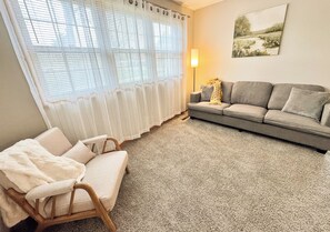 Una sala de estar con un sofá gris, una silla de madera, una lámpara y un cuadro en la pared.
