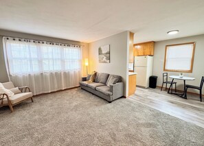 Ein Wohnzimmer mit einer Couch, einem Sessel, einem kleinen Tisch und einer Küchenzeile mit Kühlschrank und Schränken.