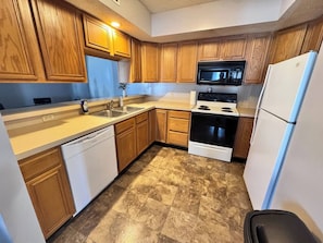 A compact kitchen with wooden cabinets, a dishwasher, a stove, a microwave, and a refrigerator. - 511 Serenity on Shugart (Beatrice)
