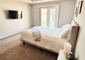 A bedroom with a bed, a television mounted on the wall, a door leading to an outdoor area, and a small bedside table with a lamp. - 511 Serenity on Shugart (Beatrice)