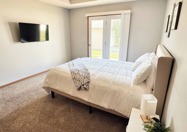 A bedroom with a bed, a television mounted on the wall, a door leading to an outdoor area, and a small bedside table with a lamp.