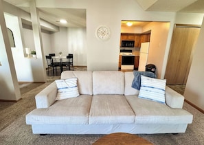 A living room with a beige sofa, two pillows, a dining area with a table and chairs, and a kitchen in the background.