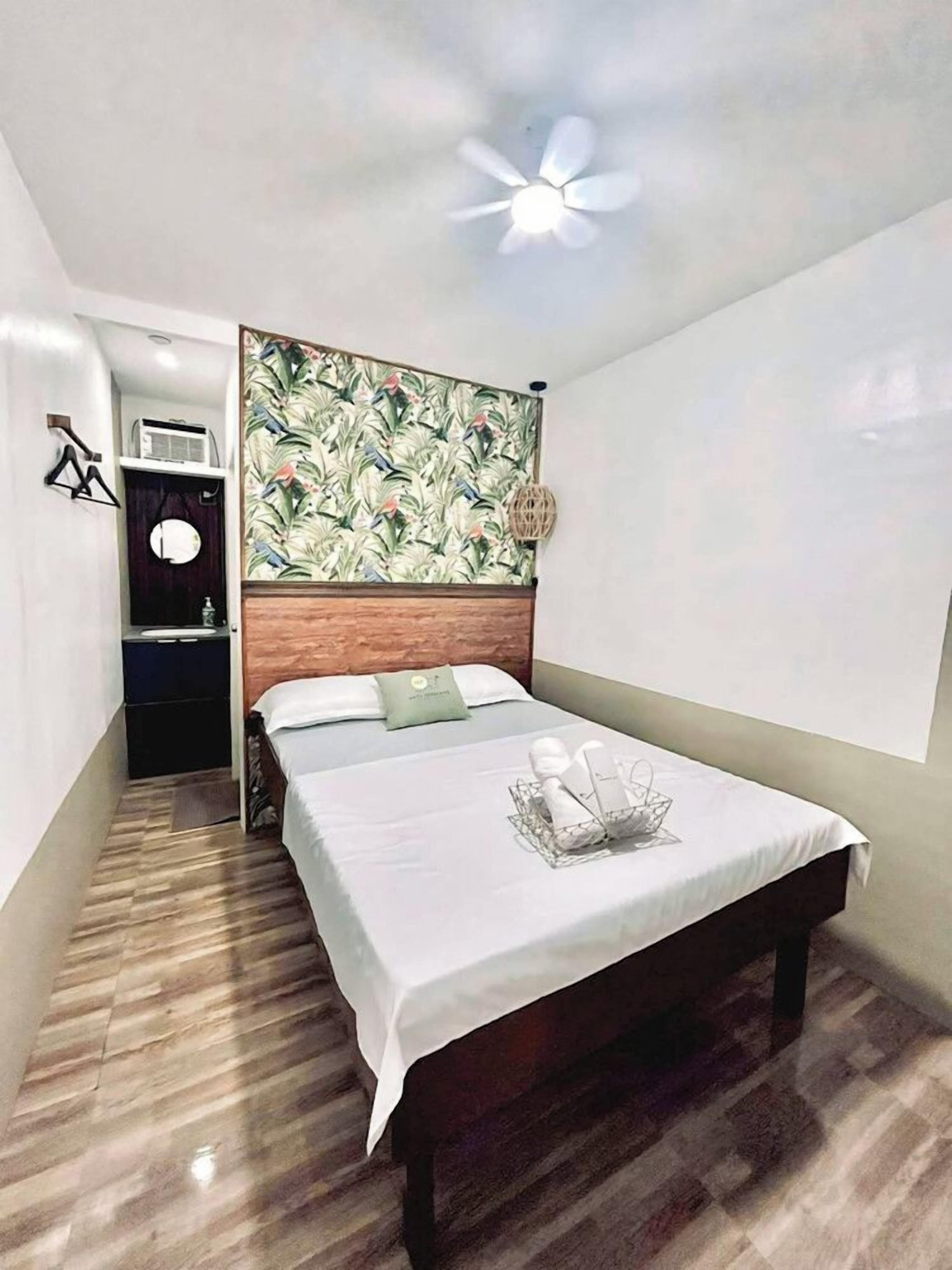 Quarto de hotel com cama, cabeceira estampada com flores, ventoinha no teto e uma mesinha de cabeceira.