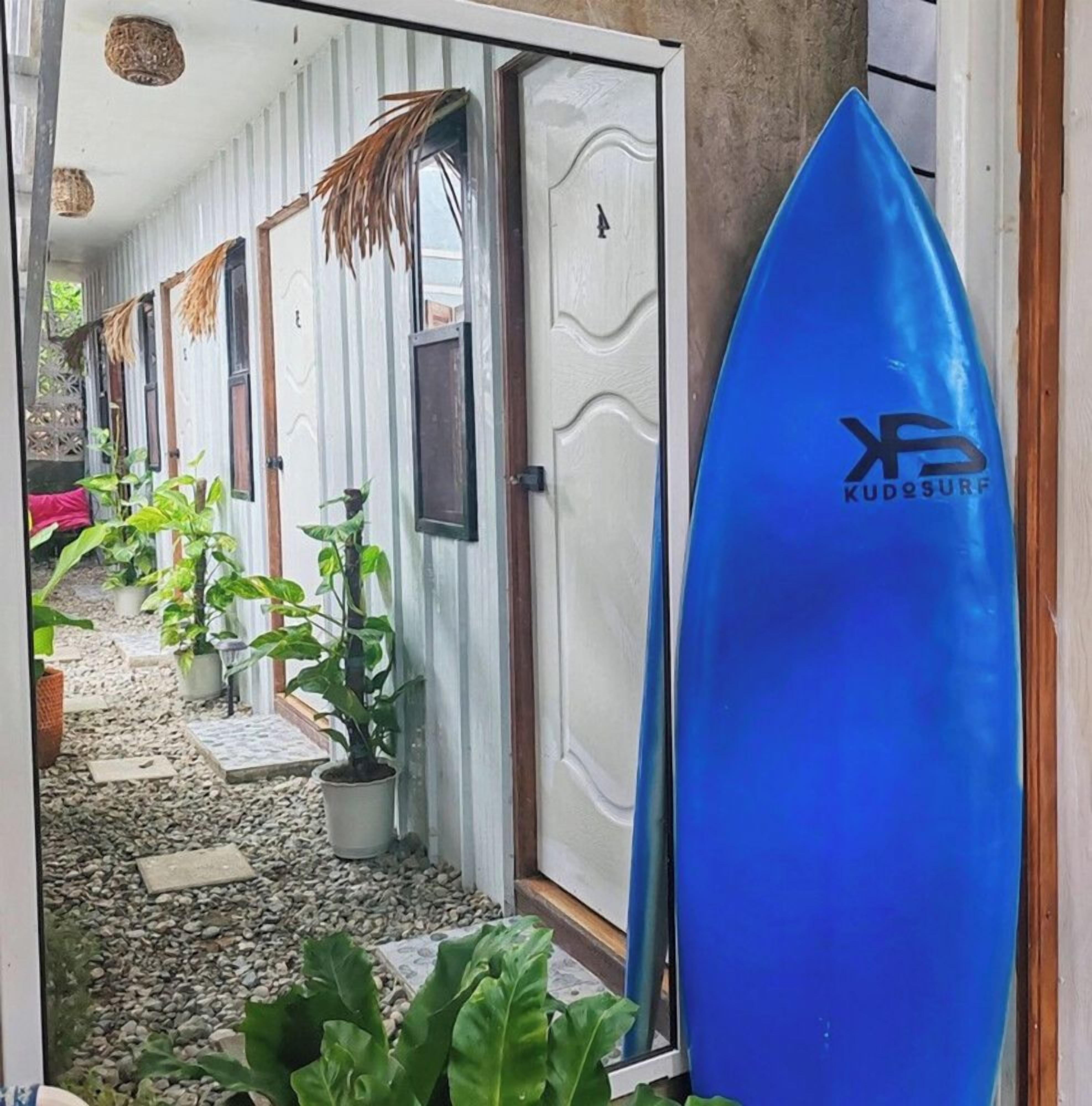 A hallway with a surfboard leaning against a door, potted plants, and a tiled floor.
