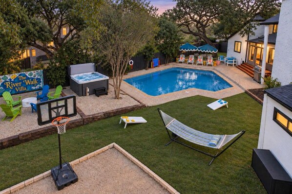 Un patio trasero con piscina, jacuzzi, aro de baloncesto, sillas de descanso y una hamaca.