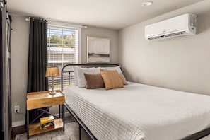 A bedroom with a bed, bedside table, lamp, air conditioning unit, and a window with curtains. - Pets Welcome! Tiny House + Guadalupe River Access (New Braunfels)