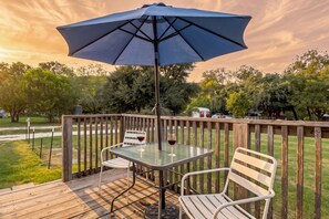 Une terrasse en bois avec une table et des chaises, un parasol, et une vue sur des arbres et un véhicule au loin.
