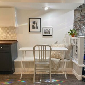 A dining area with a white table and chair, two framed pictures on the wall, a potted plant, and a stone wall. - Charming & Furnished Roncesvalles Suite. Pet Friendly! (Toronto)