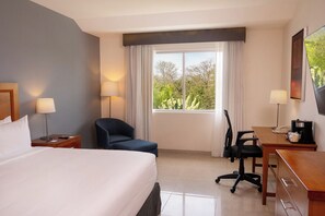 Habitación de hotel con una cama, un escritorio, una silla, una lámpara y una ventana con vista a zonas verdes.