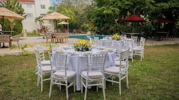 Un espacio de comedor al aire libre con mesas redondas y sillas blancas, rodeado de follaje y una piscina.