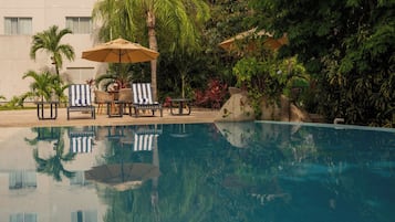 Una piscina con sillas de descanso y sombrillas, rodeada de abundante vegetación y un edificio al fondo.