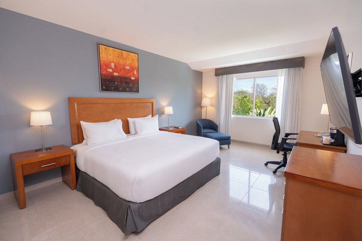 Una habitación de hotel con una cama grande, un escritorio, una silla, un televisor y una ventana con cortinas.