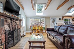 Un salon chaleureux avec une cheminée en pierre, des poutres apparentes en bois au plafond, un canapé en cuir et un coin repas avec des chaises en bois.