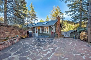 A log cabin with a stone patio, outdoor seating, and a view of surrounding trees. - Oakwood Lodge! Secluded Forest Retreat w/ Hot Tub (Green Mountain Falls)