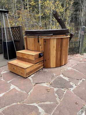 Un bain à remous avec des marches en bois et un barbecue en métal, installé sur une terrasse en pierre avec des arbres en arrière-plan.