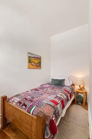 A single bed with a patterned bedspread, a small bedside table with a lamp, and a wall-mounted picture. - Charming historic bungalow in prime location (Tucson)