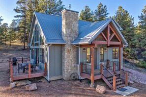 Une cabane avec une cheminée en pierre, une véranda dotée d’une balustrade en bois et une terrasse aménagée avec du mobilier d’extérieur.