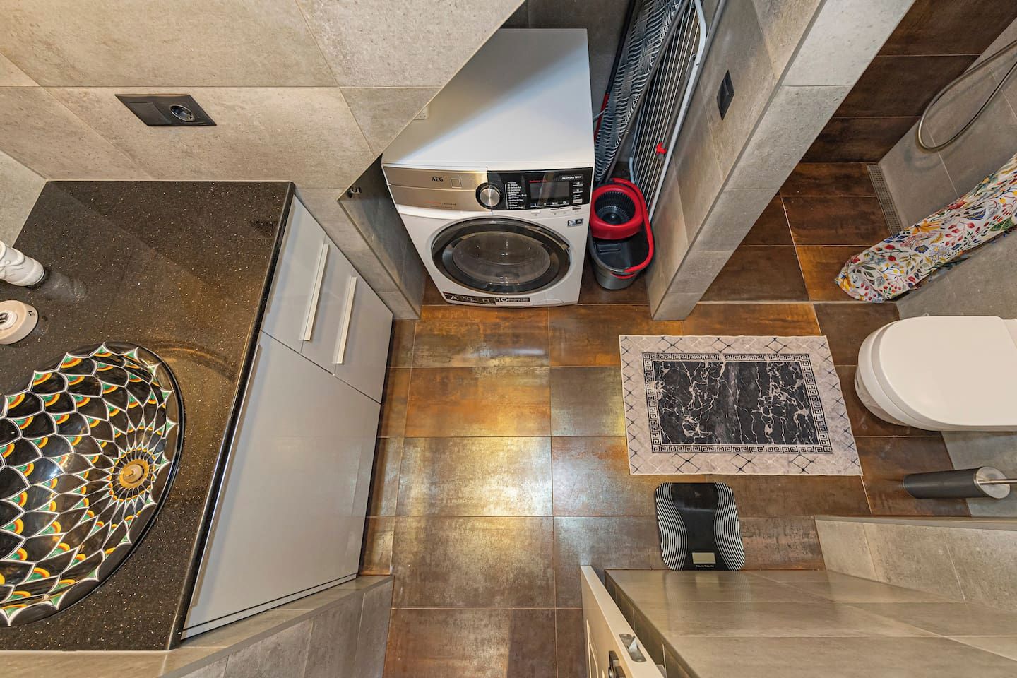 A compact bathroom with a washing machine, a toilet, and a patterned rug.