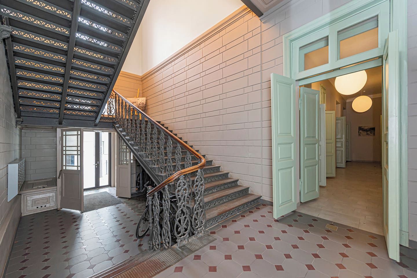 A staircase with ornate metal railings and a wooden handrail, leading to a room with a light-colored tiled floor and a green door.