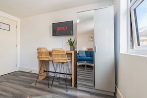Ein modernes Wohnzimmer mit einem wandmontierten Flachbildfernseher, der 'NETFLIX' anzeigt, zwei Korbstühlen, einem kleinen Holztisch, einem blauen Sofa und einer Topfpflanze.