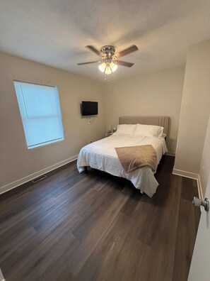 A bedroom with a bed, a ceiling fan, a TV, and a window.