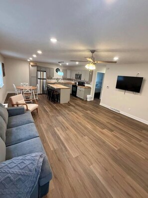 A modern living room with a sofa, a dining area with chairs, a kitchen with a central island, and a ceiling fan. - Nebraska City Nook - Oak (1310 1st Ave) (Nebraska City)