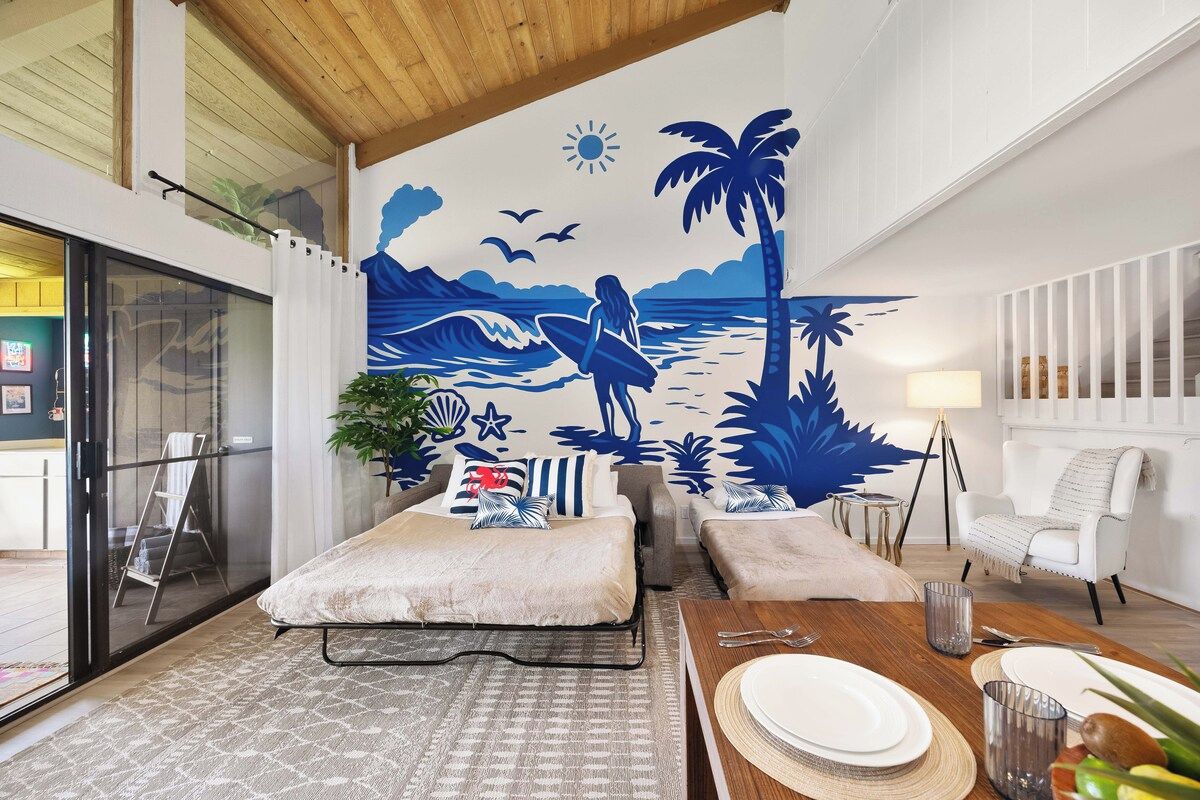 A room with a bed, a dining table set for two, a mural of a beach scene, and a white chair.