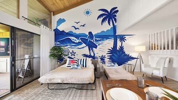 A room with a bed, a dining table set for two, a mural of a beach scene, and a white chair.
