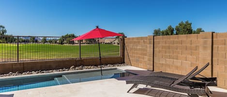 Una piscina con un paraguas rojo, sillas de descanso y vistas a un campo verde.
