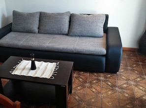 Ein graues Sofa mit mehreren Kissen, ein kleiner Tisch mit einer Flasche und einem Streifenstoff sowie geflieste Bodenbeläge mit Muster.