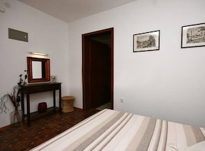 Ein Zimmer mit einem Bett, einem Holztisch mit Spiegel, einem kleinen Hocker und zwei gerahmten Bildern an der Wand.