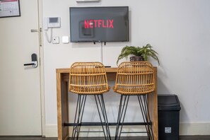 Ein Zimmer mit Schreibtisch aus Holz, zwei Korbstühlen, einem Blumentopf, einem Mülleimer und einem an der Wand montierten Fernseher, der 'NETFLIX' anzeigt.