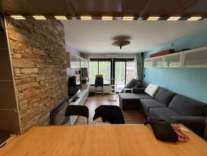 A modern living room with a stone accent wall, a sofa, a dining table, and a view through large windows. - Renovated studio, panoramic view - 4 guests (Pra-Loup)