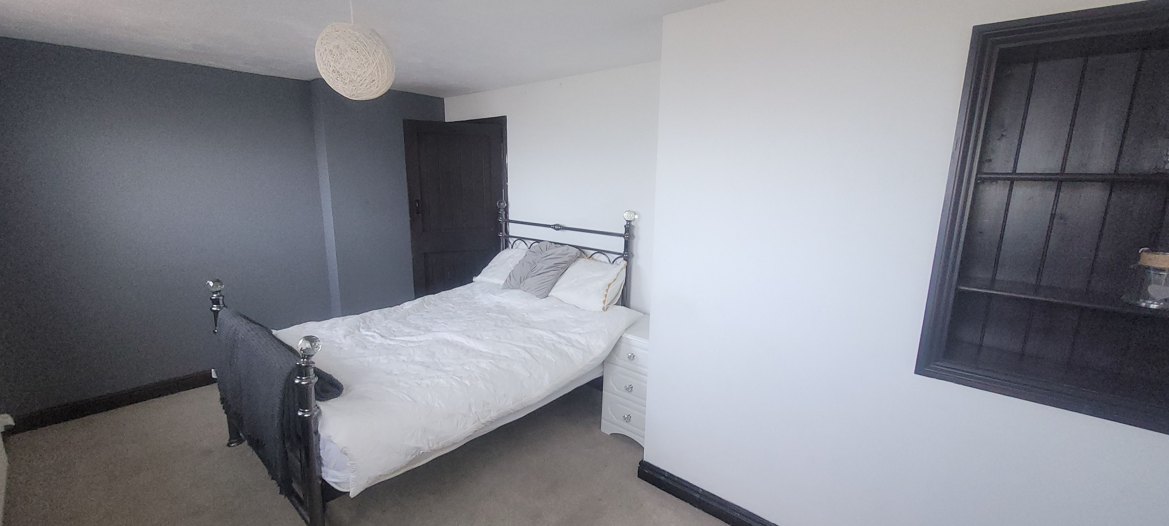 Ein Einzelbett in einem Zimmer mit dunkler Wand, weißer Decke und einem Fenster mit dunklem Rahmen.