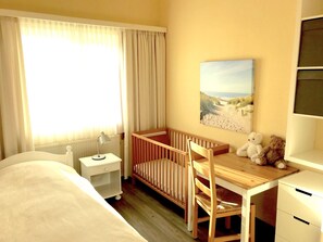 Ein Hotelzimmer mit Babybett, Schreibtisch mit Stuhl, Lampe, Teddybär und einem Bild an der Wand.