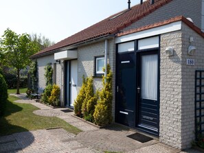 A two-story building with a brick facade, numbered 180, featuring a dark-colored door and windows, a paved driveway, and a small garden area with shrubs and a bench. - Cozy, very well-equipped vacation home for up to 5 people, within walking distance of the beach (Julianadorp)