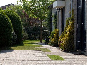 Ein gepflasterter Weg führt zu einem Gebäude mit Backsteinfassade, umgeben von gepflegten Hecken und einem Baum.