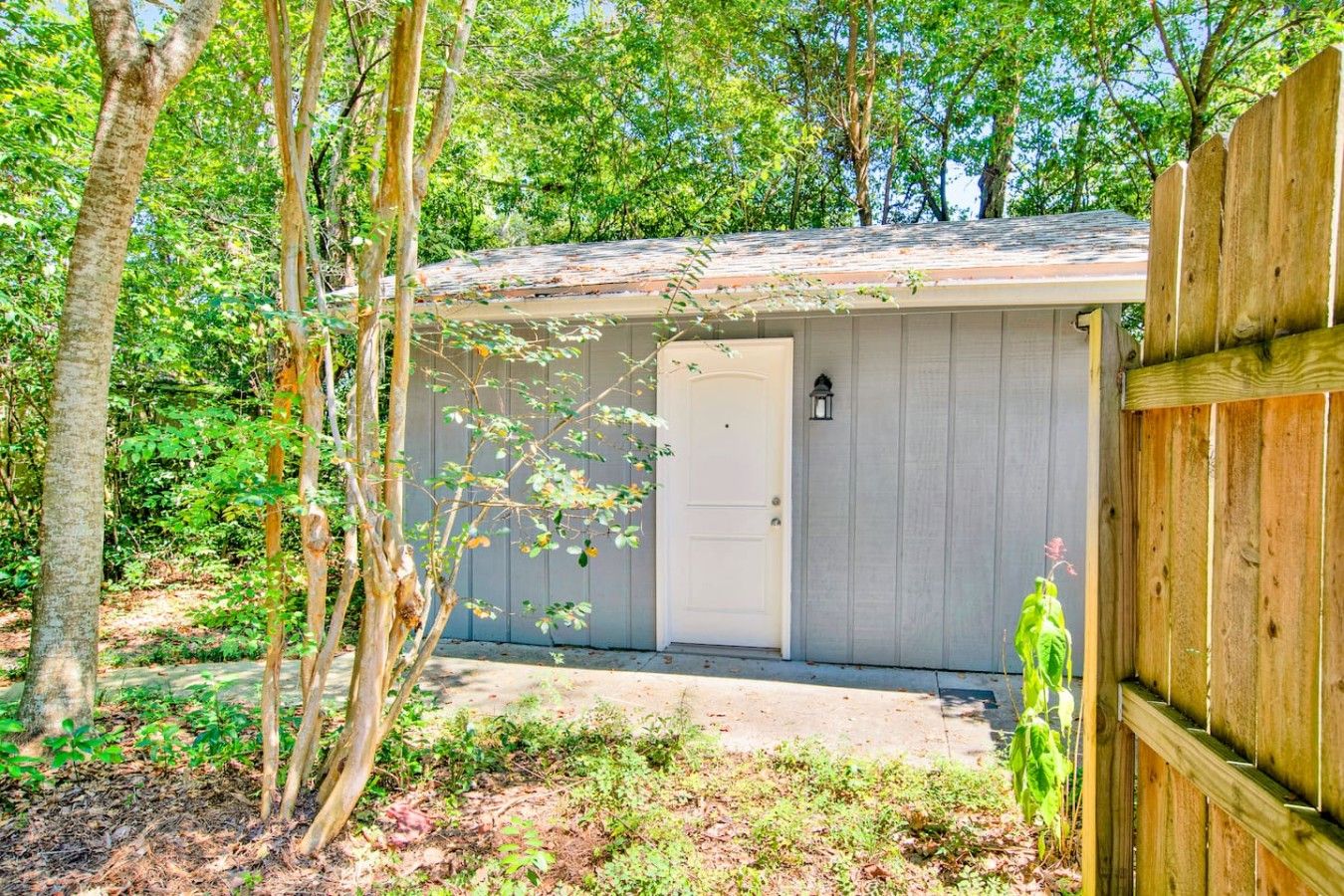 A small, single-story shed with a white door and a window, surrounded by trees and a wooden fence.