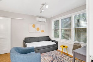 A room with a grey sofa, a blue chair, a small yellow stool, a patterned rug, and a window with blinds. - College Cottage (Mobile)