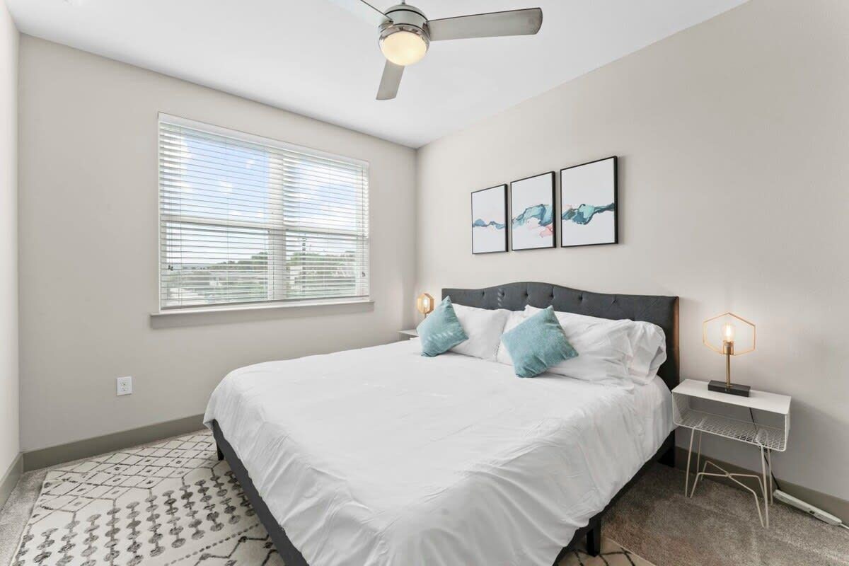 A bedroom with a bed, a ceiling fan, a bedside table with a lamp, and artwork on the wall.