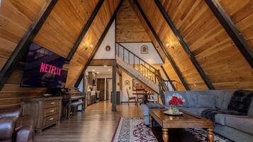 Ein Dachzimmer mit geneigtem Holzboden, einem Flachbildfernseher, einem Klavier, einer Couch und einer Treppe, die in eine weitere Ebene führt.