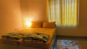 A single bed with a headboard, a bedside table with a lamp, a window with curtains, and a patterned rug on the floor.