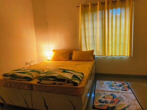 Un lit simple avec une tête de lit, une table de chevet avec une lampe, une fenêtre avec des rideaux et un tapis à motifs sur le sol.