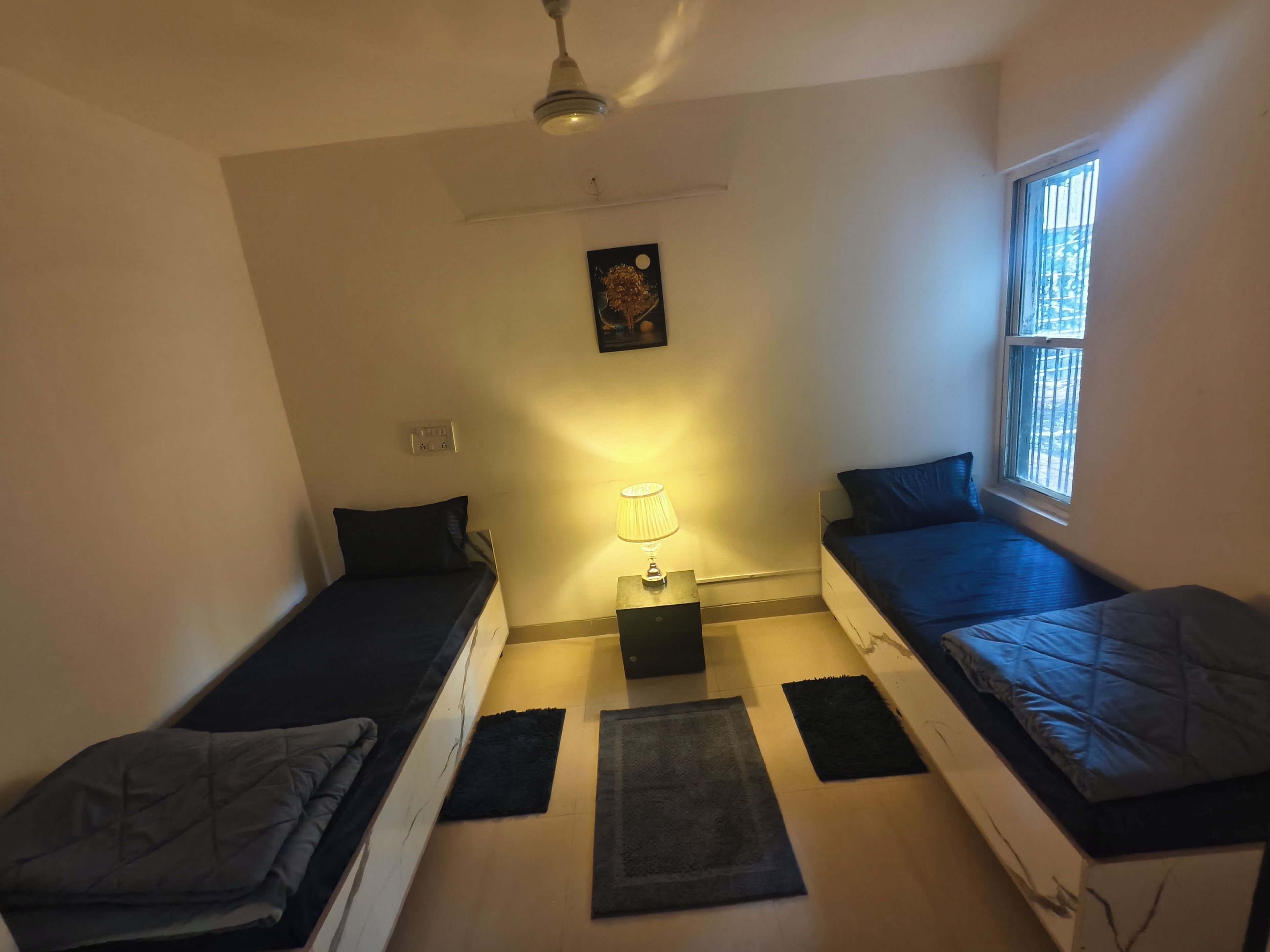 A small, compact room with two single beds, a ceiling fan, a window with bars, and a wall-mounted light fixture.