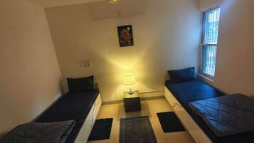 A small, compact room with two single beds, a ceiling fan, a window with bars, and a wall-mounted light fixture.