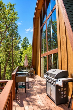 Una terraza de madera con una parrilla y mobiliario exterior, rodeada de árboles y un ventanal grande.