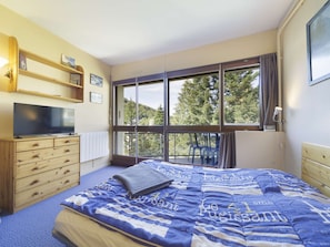 Een slaapkamer met een bed, een televisie op een houten dressoir, een raam met uitzicht op bomen en een plank met spullen.