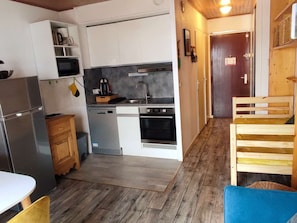 A compact kitchen with a refrigerator, microwave, sink, and oven. There is a wooden bench and a small table. - Ideal studio in the center - 3 guests (Valloire)