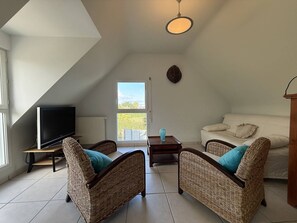 A loft-style living room with a television, two wicker chairs, a white sofa, and a view through a window. - Quiet house with WIFI 600m from the sea (Trégastel)