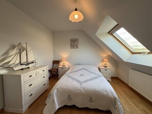 A bedroom with a sloped ceiling, a skylight, a bed with a quilted cover, a dresser, a chair, and a model ship. - Quiet house with WIFI 600m from the sea (Trégastel)